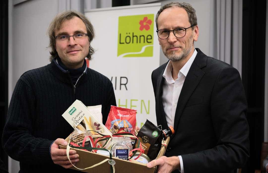 Stefan Galle und Bernd Poggemöller halten zusammen einen Präsentkorb. Im Korb sind verschiedene Snacks und Süßigkeiten. Beide lächeln.