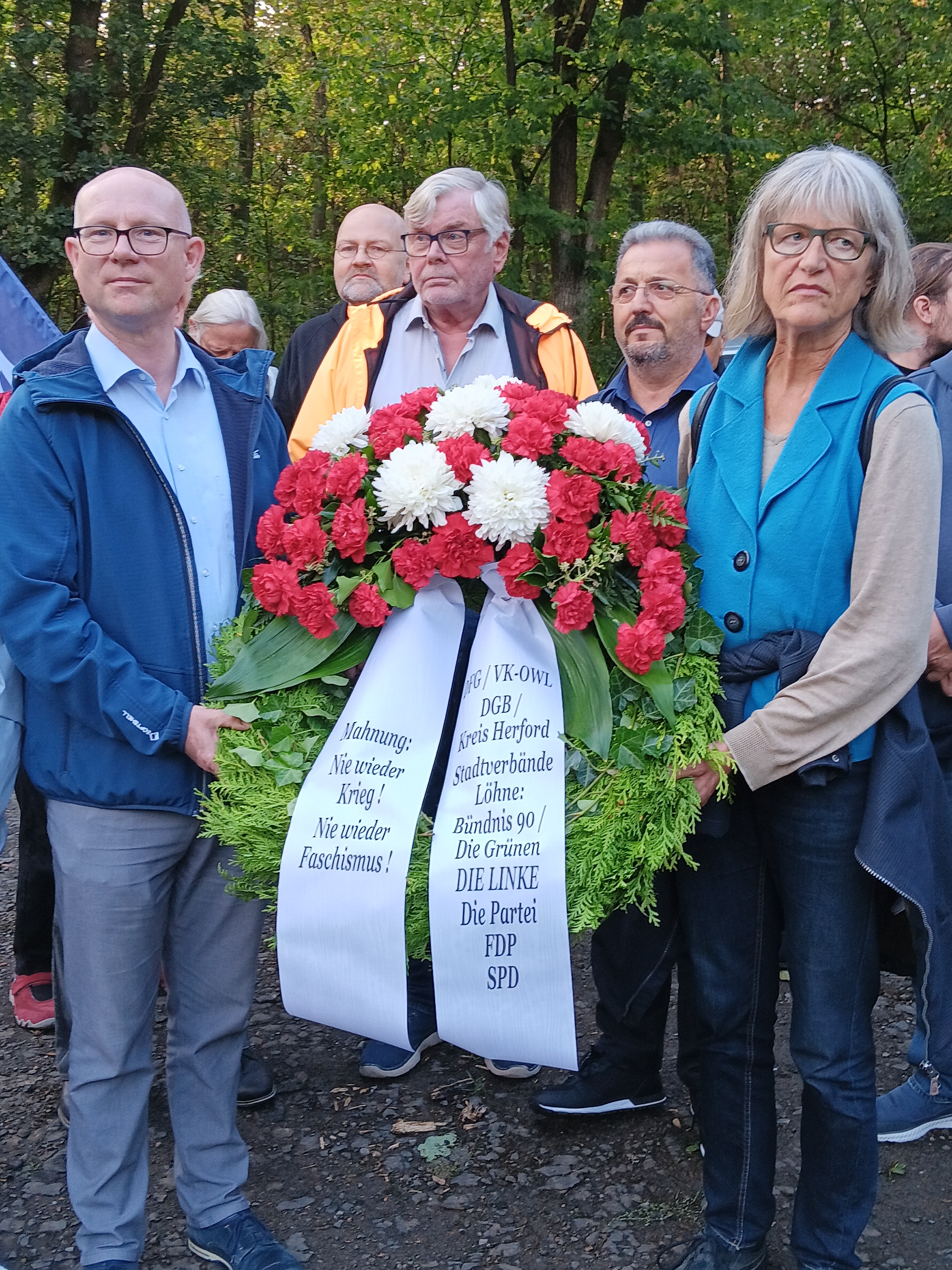 Teilnehmer präsentieren Kranz. Widmung: Mahnung: Nie wieder Krieg! Nie wieder Faschismus! DFG / VK-OWL DGB / Kreis Herford Stadtverbände Löhne: Bündnis 90 DIE GRÜNEN / DIE LINKE Die Partei FDP SPD